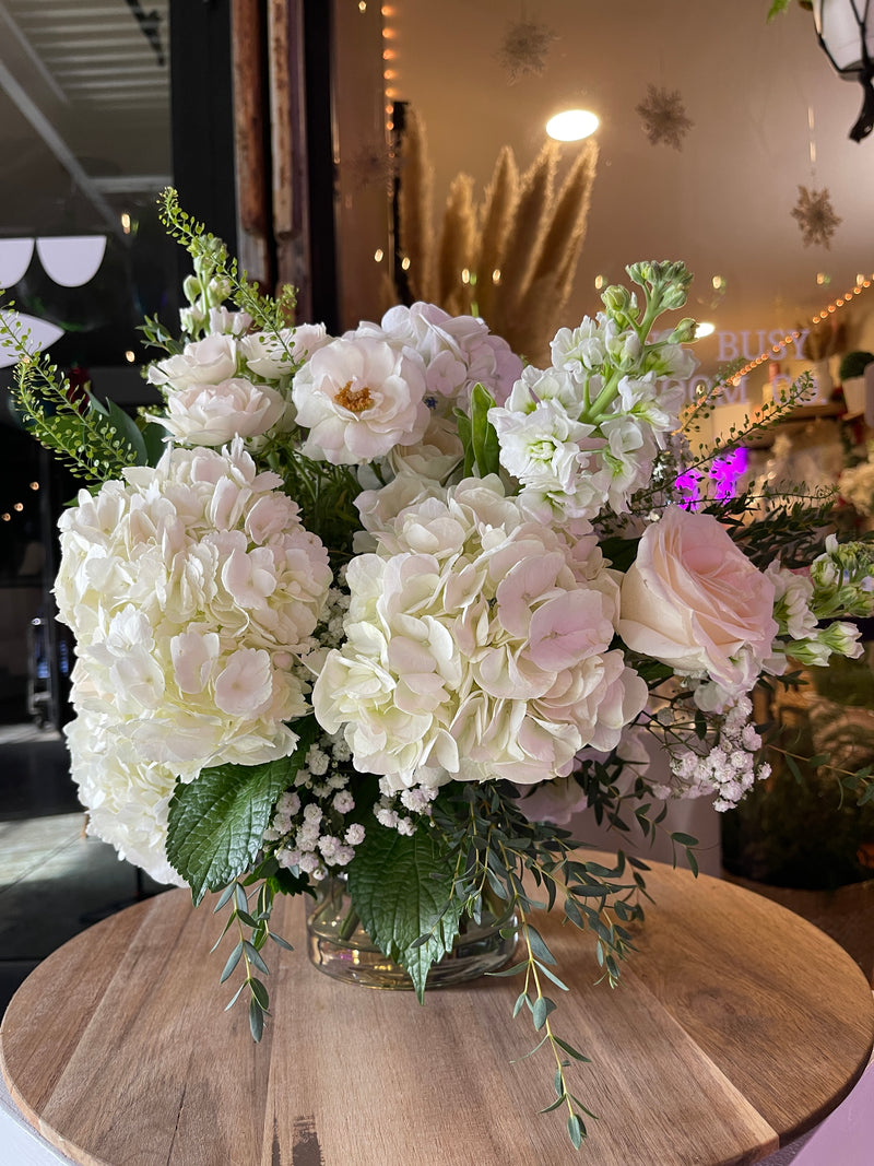 A white centerpiece featuring roses, hydrangea, stock, and eucalyptus, arranged in a clear glass vase on a wooden table.